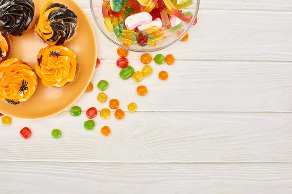 Vue de dessus des bonbons gommeux colorés, des bonbons et des cupcakes sur la table en bois blanche, festin d'Halloween — Photo de stock