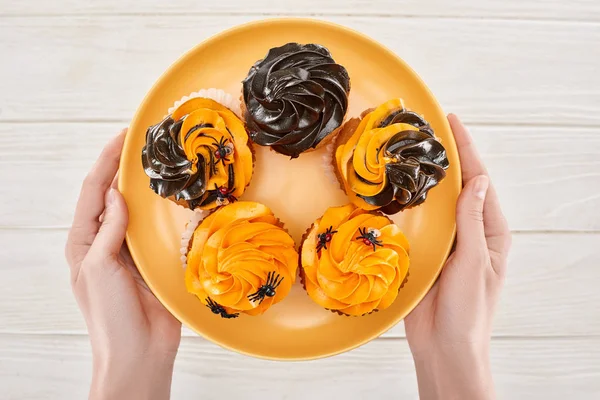 Vista parziale della donna che tiene deliziosi cupcake con ragni sul piatto arancione sul tavolo di legno bianco, Halloween trattare — Foto stock