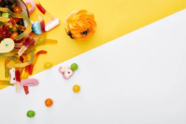 Draufsicht auf bunte Gummiwürmer in der Nähe von Cupcake auf weißem und gelbem Hintergrund, Halloween-Leckerbissen — Stockfoto