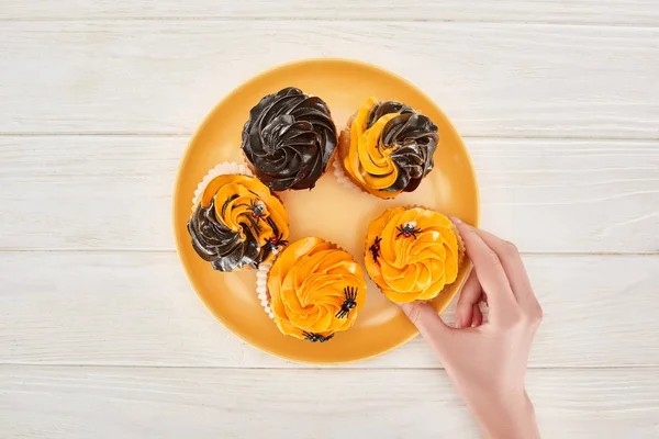 Abgeschnittene Ansicht einer Frau, die köstlichen Cupcake mit Spinnen von orangefarbenem Teller auf weißem Holztisch nimmt, Halloween-Leckerbissen — Stockfoto