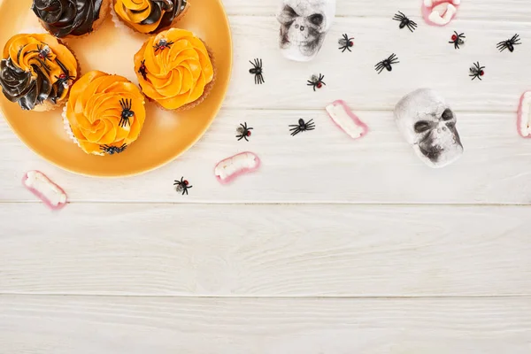 Vista superior de cupcakes em prato laranja, dentes de goma, crânios e aranhas em mesa de madeira branca, deleite de Halloween — Fotografia de Stock