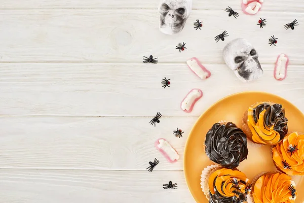 Vista superior de cupcakes en plato naranja, dientes gomosos, cráneos y arañas en mesa de madera blanca, regalo de Halloween - foto de stock