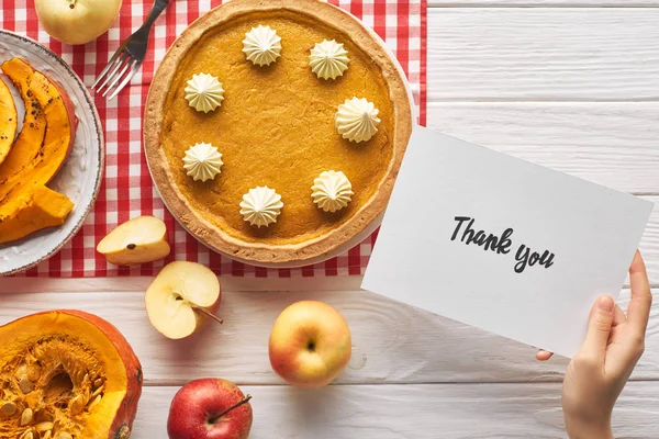 Обрізаний вид жінки, що тримає подяку вам листівку біля гарбузового пирога на дерев'яному білому столі з яблуками — стокове фото