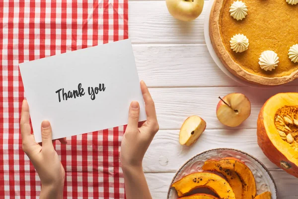Vue partielle de la femme tenant carte de remerciement près de tarte à la citrouille sur table blanche en bois avec des pommes — Photo de stock