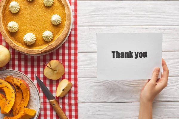 Vue recadrée de femme tenant carte de remerciement près de tarte à la citrouille sucrée sur table blanche en bois avec des pommes — Photo de stock