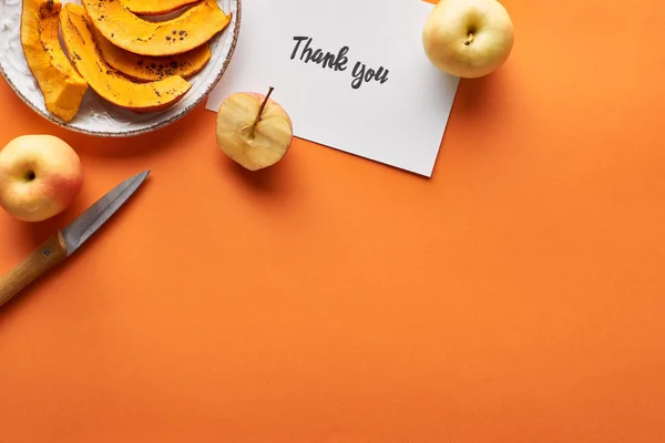 Vista superior de calabaza, manzanas, cuchillo y tarjeta de agradecimiento sobre fondo naranja - foto de stock