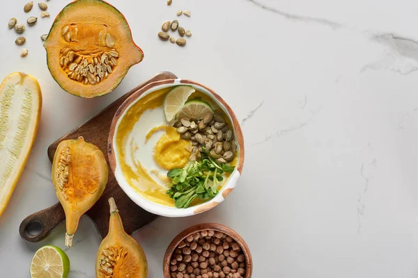 Top view of autumnal mashed pumpkin soup in bowl on wooden cutting board near pumpkin, lime and chickpea on marble surface — Stock Photo