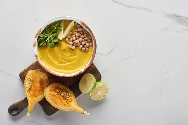 Vista superior de la sopa de calabaza molida otoñal en un tazón en una tabla de cortar de madera cerca de la calabaza, cal en la superficie de mármol - foto de stock