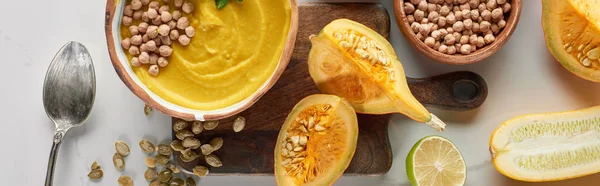 Top view of autumnal mashed pumpkin soup in bowl on wooden cutting board near pumpkin, lime and chickpea on marble surface, panoramic shot — Stock Photo