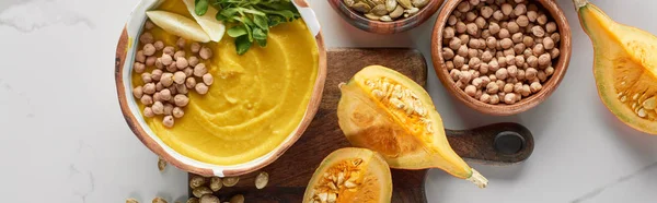 Top view of autumnal mashed pumpkin soup in bowl on wooden cutting board near pumpkin, chickpea on marble surface, panoramic shot — Stock Photo