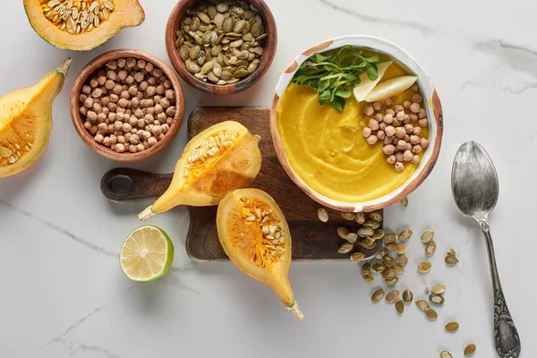 Top view of autumnal mashed pumpkin soup in bowl on wooden cutting board near pumpkin, seeds and chickpea on marble surface — Stock Photo