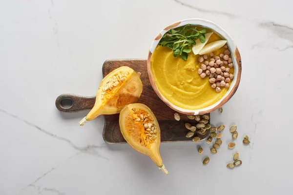 Top view of autumnal mashed pumpkin soup in bowl on wooden cutting board near pumpkin, seeds on marble surface — Stock Photo