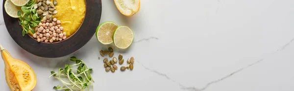 Vista superior de la sopa de calabaza molida otoñal en un tazón cerca de semillas de calabaza, brotes y cal en la superficie de mármol, plano panorámico - foto de stock