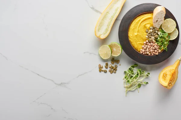 Vista dall'alto della zuppa autunnale di zucca in ciotola vicino a zucchine, semi di zucca, germogli e lime sulla superficie di marmo — Foto stock