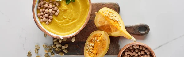 Top view of autumnal mashed pumpkin soup in bowl on wooden cutting board near pumpkin, seeds and chickpea on marble surface — Stock Photo