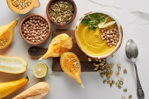 Vista superior de la sopa de calabaza molida otoñal en un tazón en una tabla de cortar de madera cerca de los ingredientes en la superficie de mármol - foto de stock