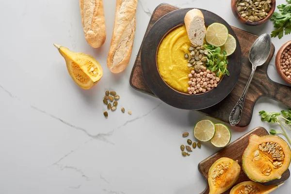 Top view of autumnal mashed pumpkin soup in bowl on wooden cutting board near pumpkin, seeds, lime and bread on marble surface — Stock Photo