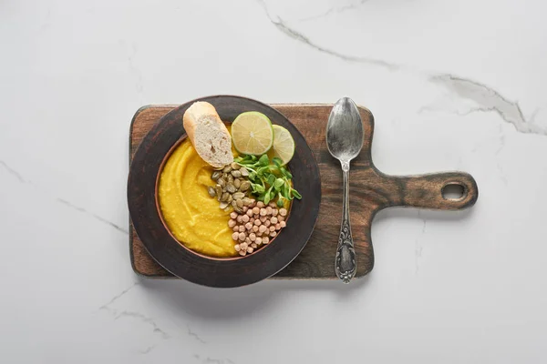 Top view of autumnal mashed pumpkin soup in bowl on wooden cutting board with spoon on marble surface — Stock Photo