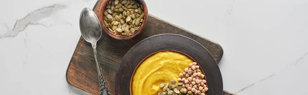 Panoramic shot of autumnal mashed pumpkin soup in bowl on wooden cutting board with spoon and seeds on marble surface — Stock Photo
