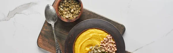 Plan panoramique de soupe à la citrouille en purée automnale dans un bol sur une planche à découper en bois avec cuillère et graines à la surface du marbre — Photo de stock