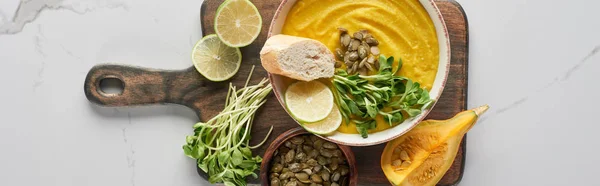 Tiro panorâmico de deliciosa sopa de abóbora purê na placa de corte de madeira na superfície de mármore com ingredientes — Fotografia de Stock