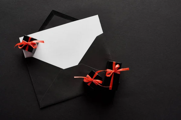 Vista superior do envelope preto com cartão em branco perto de caixas de presente decorativas no fundo preto — Fotografia de Stock