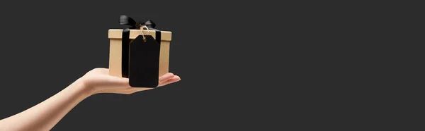 Vista cortada de mulher segurando caixa de presente com etiqueta em branco preto na mão isolado em preto, tiro panorâmico — Fotografia de Stock