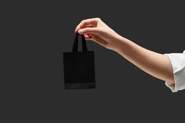 Vista ritagliata della donna che tiene in mano la piccola shopping bag nera isolata sul nero — Foto stock