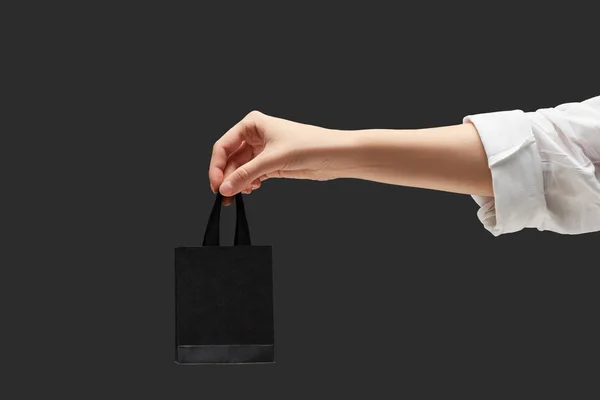 Vista recortada de la mujer sosteniendo pequeño bolso de compras negro en mano aislado en negro - foto de stock
