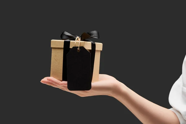 Vista recortada de la mujer sosteniendo caja de regalo con etiqueta en blanco negro en la mano aislado en negro - foto de stock