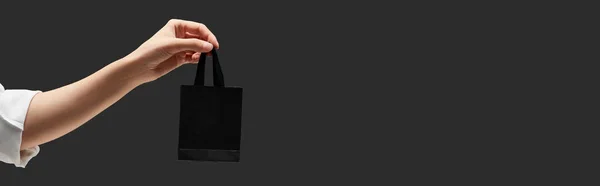 Recortado vista de la mujer sosteniendo pequeña bolsa de compras negro en mano aislado en negro, tiro panorámico - foto de stock