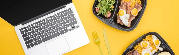 Colpo panoramico di confezioni ecologiche con verdure, carne, uova fritte e insalata, laptop, forchetta e coltello in plastica isolati su giallo — Foto stock