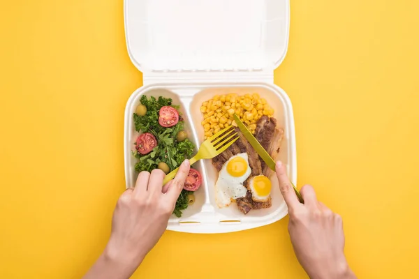 Vista recortada de la mujer comiendo del paquete ecológico con maíz, carne, huevos fritos y ensalada aislada en amarillo - foto de stock