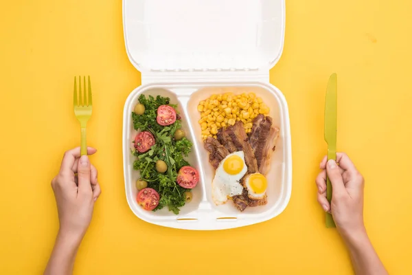 Vista recortada de la mujer sosteniendo tenedor de plástico, cuchillo y paquete ecológico con maíz, carne, huevos fritos y ensalada aislada en amarillo - foto de stock