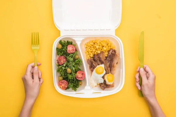 Vista ritagliata di donna in possesso di forchetta di plastica, coltello e pacchetto eco con mais, carne, uova fritte e insalata isolata su giallo — Foto stock