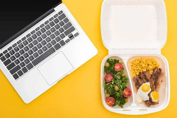 Vue du dessus de l'ordinateur portable et de l'emballage écologique avec du maïs, de la viande, des œufs frits et de la salade isolés sur jaune — Photo de stock