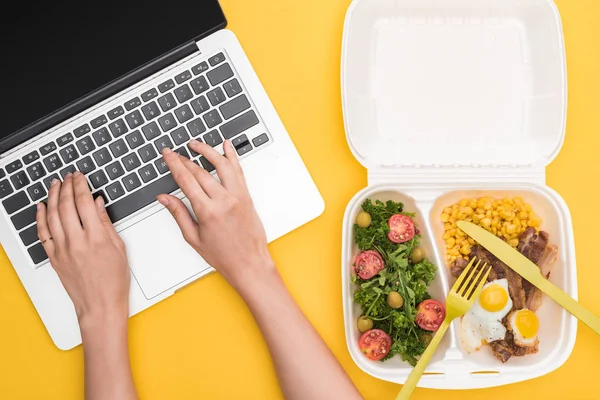 Vista recortada de la mujer usando portátil y paquete ecológico con maíz, carne, huevos fritos y ensalada aislada en amarillo - foto de stock