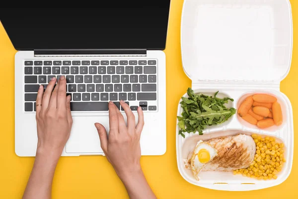 Vista cortada da mulher usando laptop e pacote ecológico com legumes, carne, ovo frito e rúcula isolada em amarelo — Fotografia de Stock