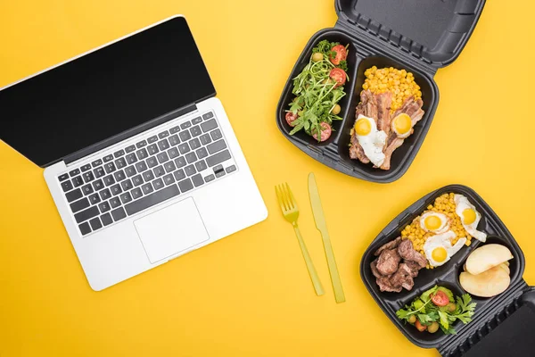 Vista superior de la computadora portátil, paquetes ecológicos con manzanas, verduras, carne, huevos fritos y ensaladas aisladas en amarillo - foto de stock