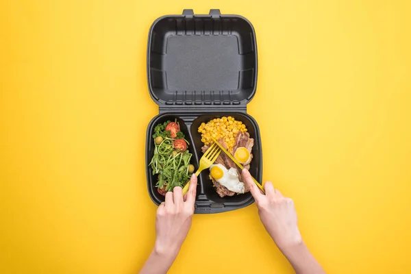 Vista recortada de la mujer comiendo del paquete ecológico con maíz, carne, huevos fritos y ensalada aislada en amarillo - foto de stock