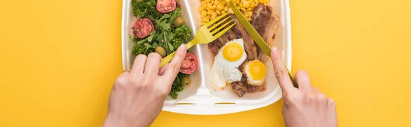 Plano panorámico de mujer comiendo de eco paquete con maíz, carne, huevos fritos y ensalada aislada en amarillo - foto de stock