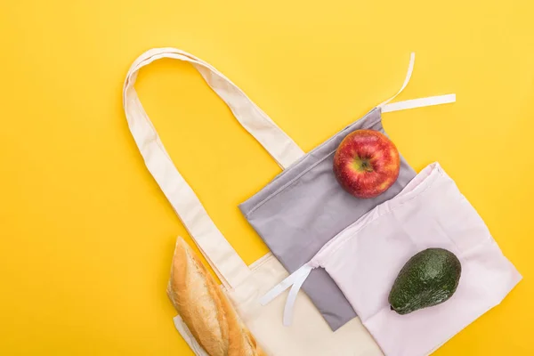 Vista superior de manzana madura, baguette y aguacate cerca de bolsas ecológicas aisladas en amarillo - foto de stock