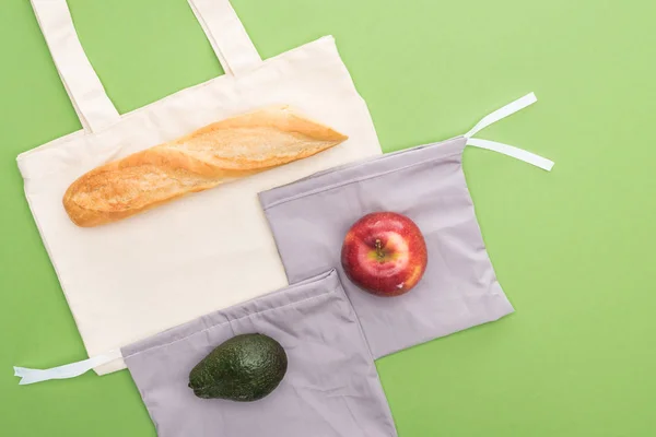 Vista superior de baguette, manzana y aguacate en bolsas ecológicas aisladas en verde - foto de stock