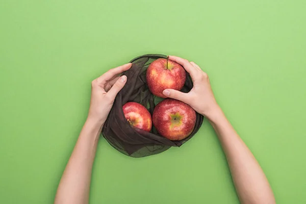 Vista parcial de la mujer sosteniendo manzanas rojas maduras en bolsa ecológica aislada en verde - foto de stock