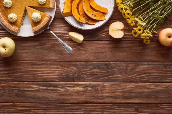 Deliziosa torta di zucca tagliata con panna montata vicino alla zucca cotta a fette, fiori gialli, mele intere e tagliate sul tavolo di legno marrone — Foto stock