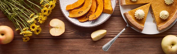 Colpo panoramico di deliziosa torta di zucca con panna montata vicino alla zucca affettata al forno, fiori gialli, mele intere e tagliate sul tavolo di legno — Foto stock