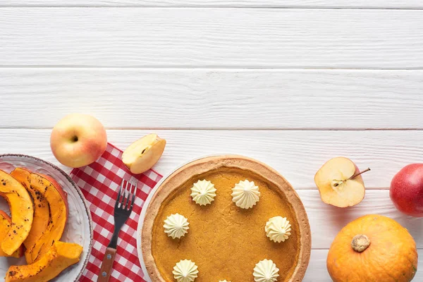 Tasty pumpkin pie with whipped cream on checkered napkin near baked and raw pumpkins, fork, whole and cut apples on white wooden table — Stock Photo