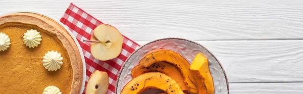 Colpo panoramico di gustosa torta di zucca con panna montata su tovagliolo a quadretti vicino alla zucca cotta a fette e mele tagliate su tavolo di legno bianco — Foto stock