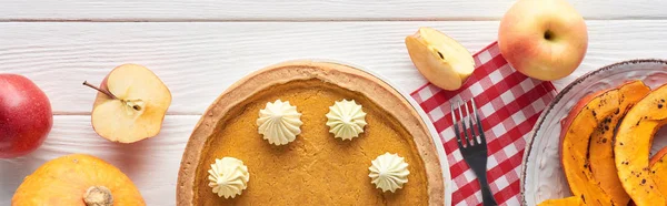 Colpo panoramico di torta di zucca con panna montata su tovagliolo a quadretti vicino a zucche cotte e crude, forchetta, mele intere e tagliate su tavolo di legno bianco — Foto stock