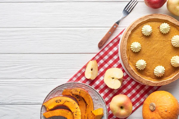 Tasty pumpkin pie with whipped cream on checkered napkin near raw and sliced baked pumpkins, cut and whole apples, and fork on white wooden table — Stock Photo